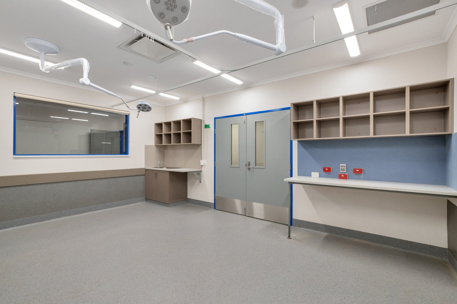 Empty operating room with surgical lights, stainless steel doors, and blue and beige walls.