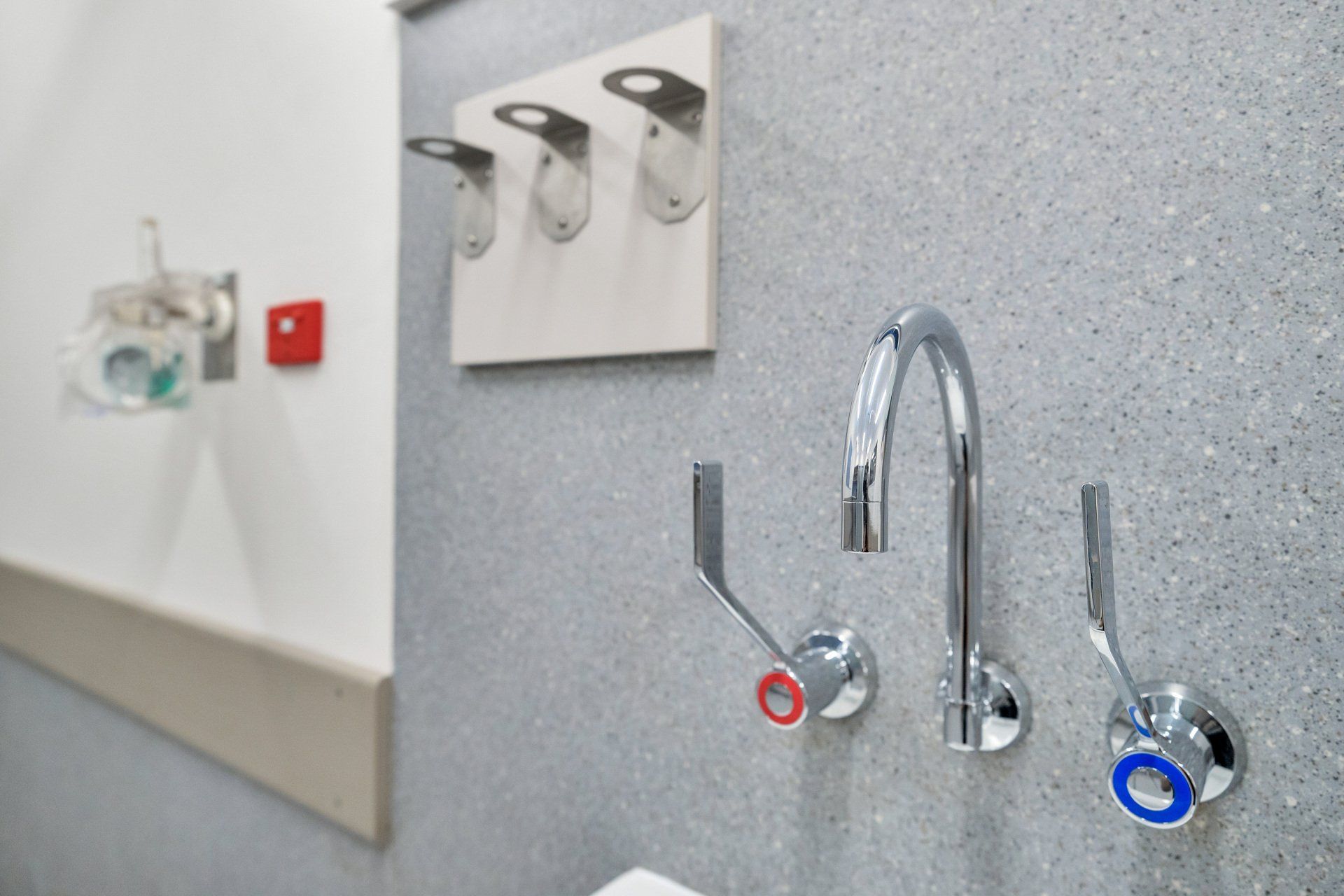 Chrome faucet with lever handles, blue/red indicators on a gray wall. Hooks and emergency button on the white wall.