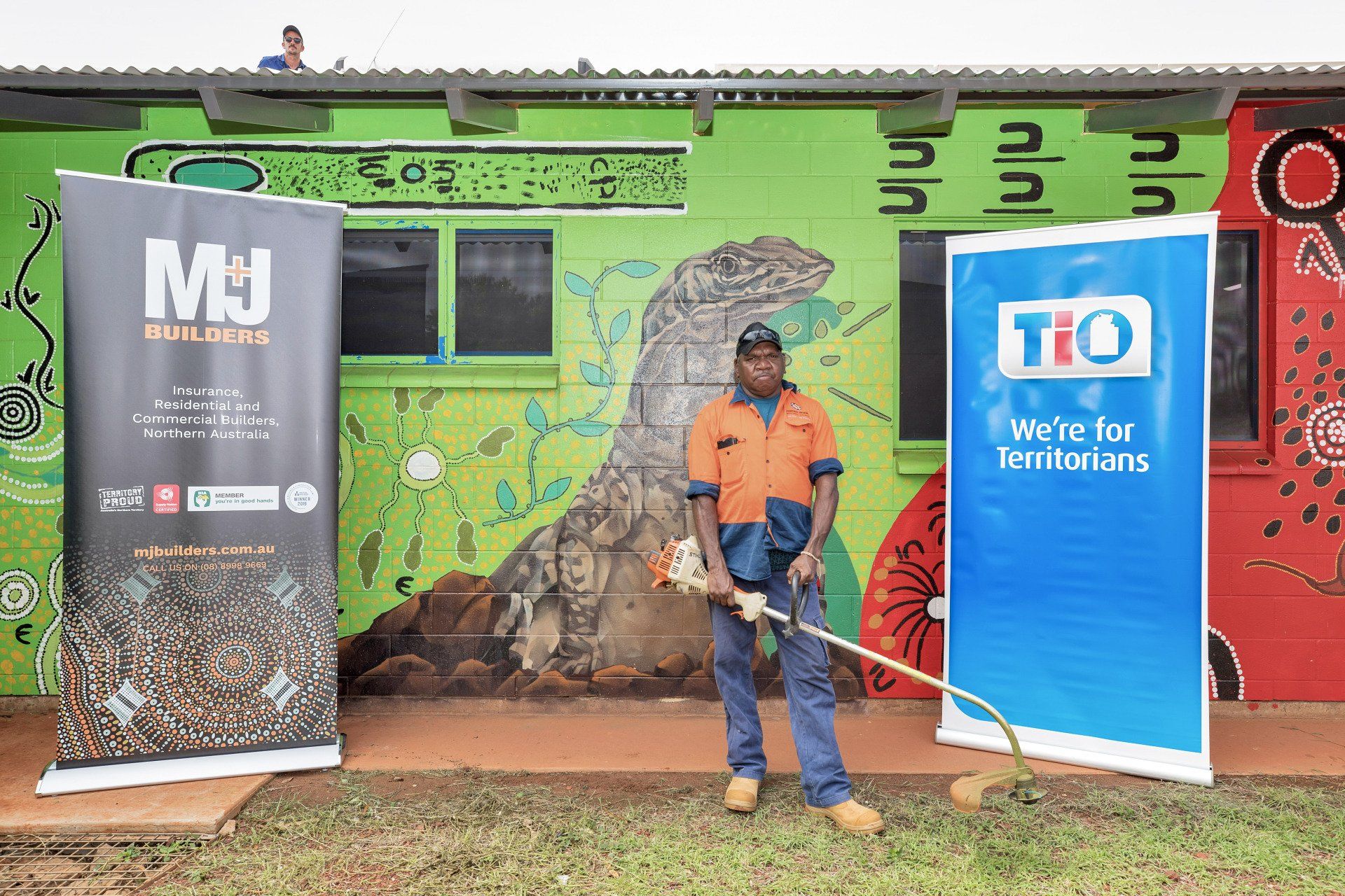 Man using weed wacker, in front of building with mural and company banners.