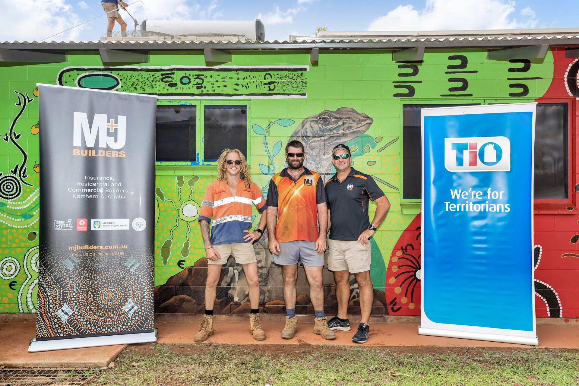 Three men stand in front of a painted wall with banners for MJJ Builders and TIO, in an outdoor setting.