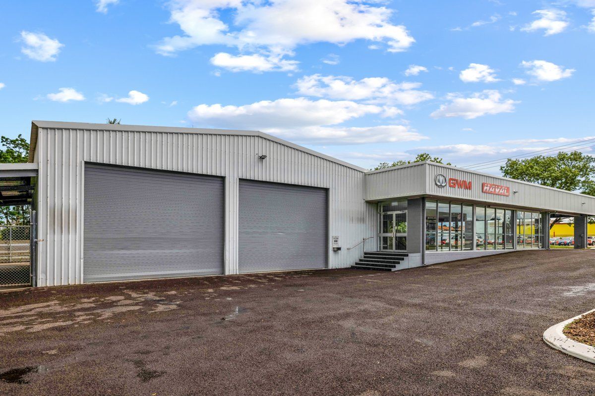 Warehouse-style building with gray roller doors, glass windows, and a paved driveway under a partly cloudy sky.