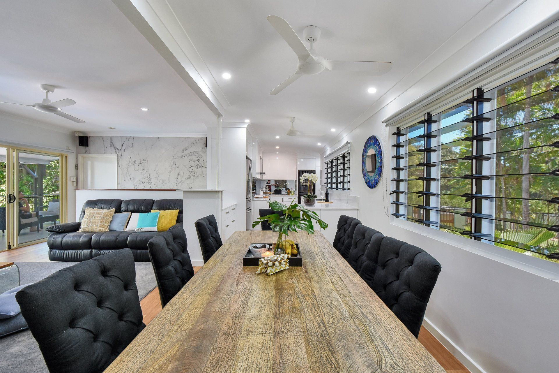 Dining room with long wooden table, black button-tufted chairs, open to living area with black sofa.