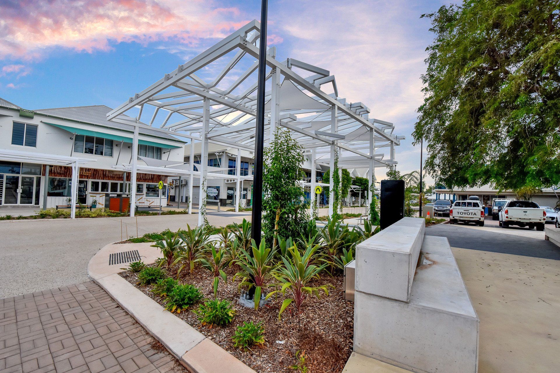 White pergola over a walkway lined with shops; a landscaped area in the foreground.