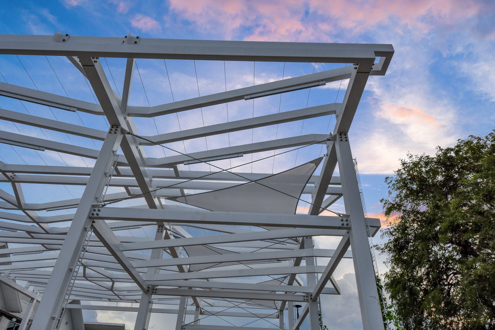 White pergola against a blue and pink sky, with a small white shade sail and tree in the corner.