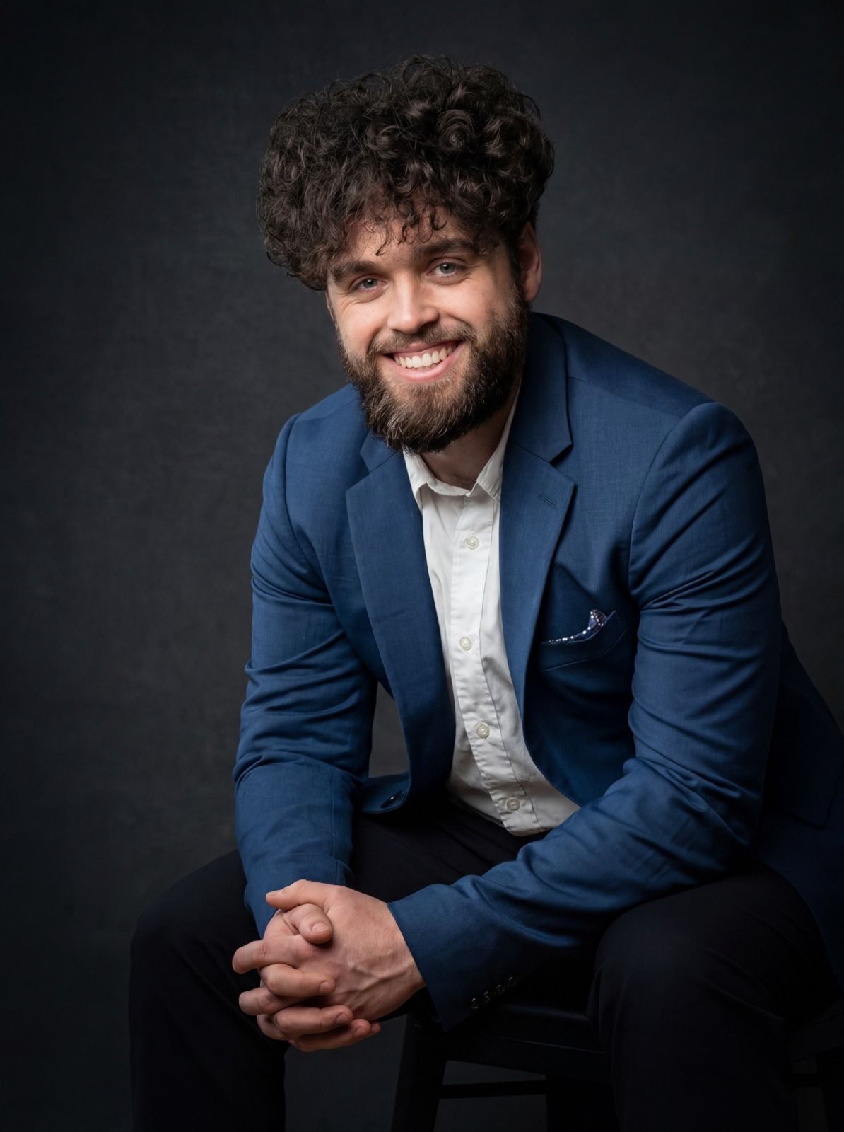 Smiling man in a blue suit seated against a dark background, hands clasped.
