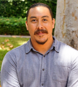 A man with a beard wearing a grey shirt is standing in front of a tree.