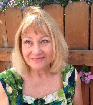 A woman in a green and blue dress is smiling in front of a wooden fence.