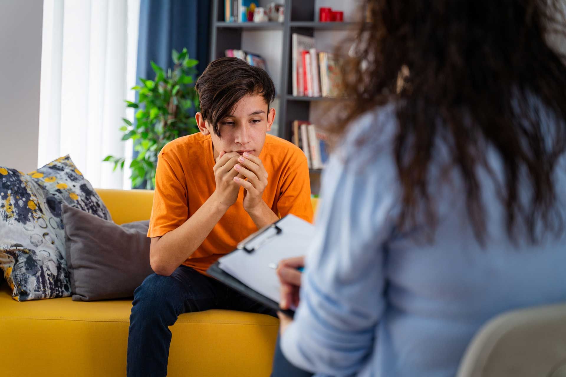 A young boy is sitting on a yellow couch talking to a woman.