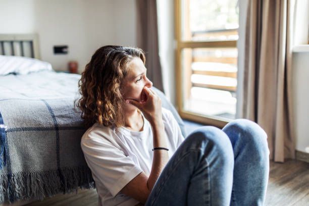 A woman is sitting on the floor in a bedroom looking out the window.