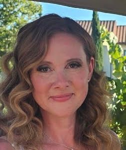 Woman with wavy blonde hair, smiling, outdoors in front of foliage and building.