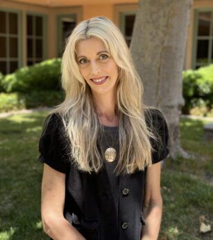 A woman with blonde hair and a black shirt is standing in front of a tree.