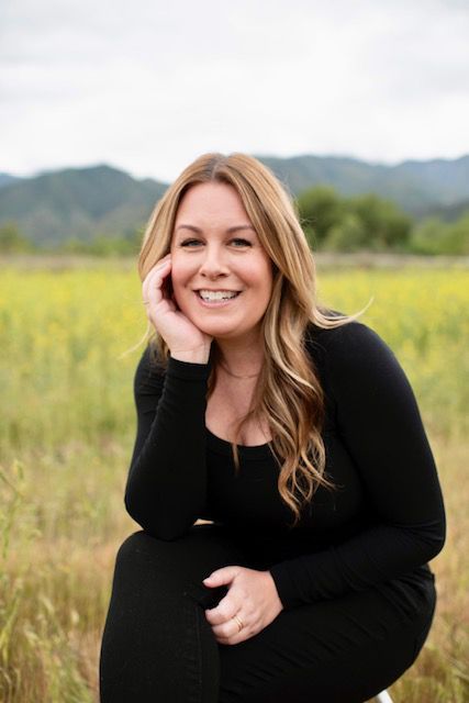 Woman with blonde hair smiles, resting her head on her hand, outdoors in a field wearing black.