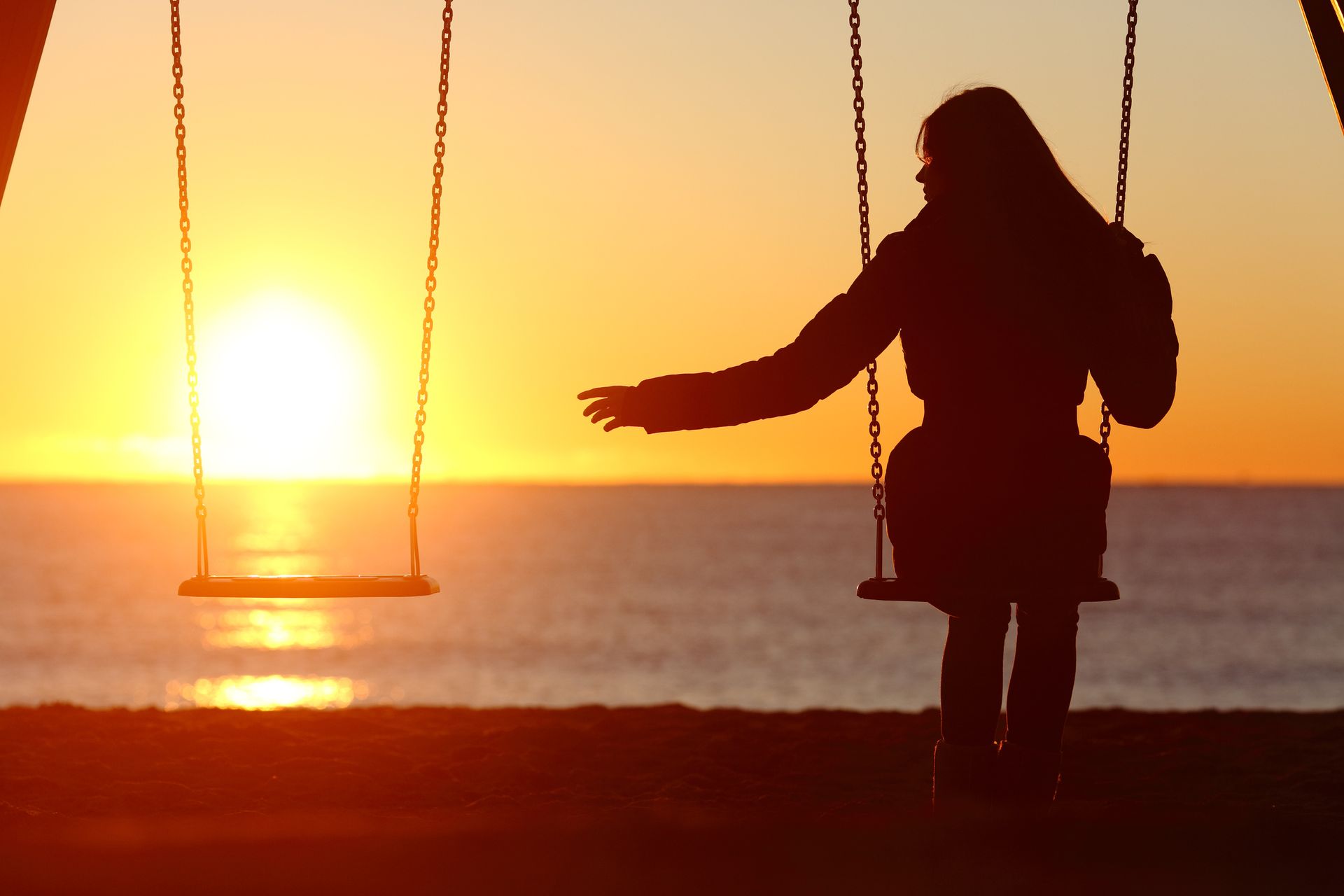 A woman is sitting on a swing in front of the ocean at sunset