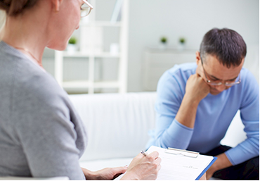 A man is sitting on a couch talking to a woman who is writing on a clipboard.