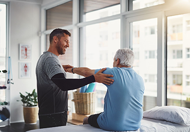 A man is helping an older man with his shoulder while sitting on a bed.
