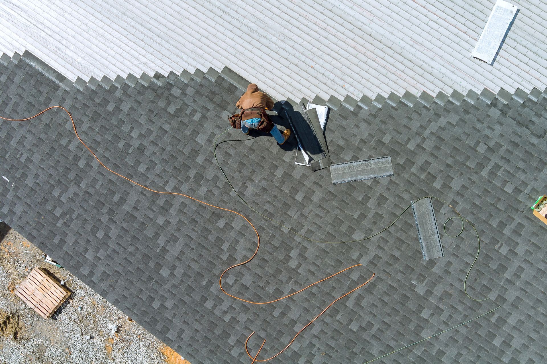 Roofer installing dark gray shingles on a roof. Aerial view shows worker, tools, and materials.