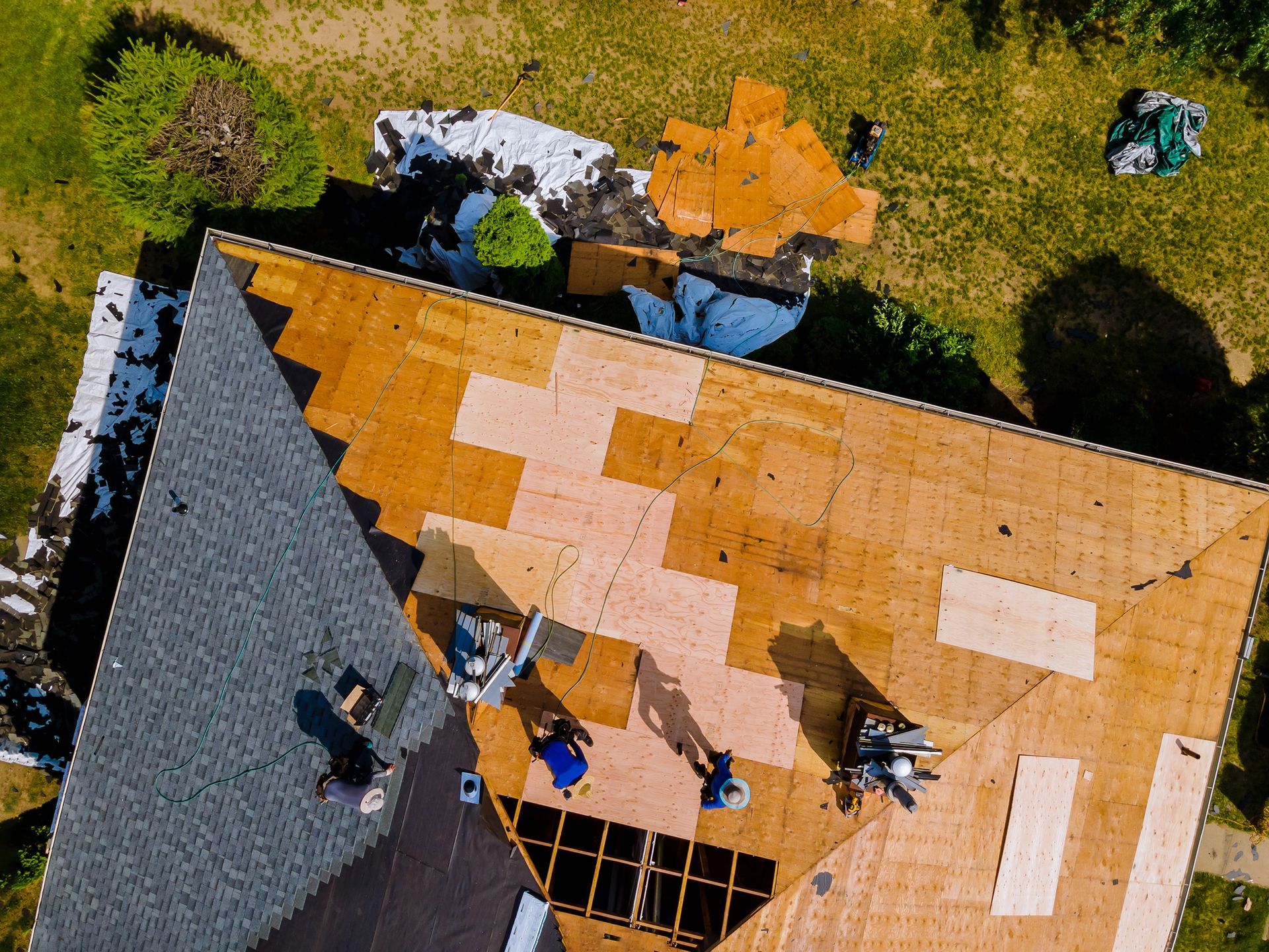 Roofers working on a house roof replacement. Wood and gray shingles visible; tools and debris on the roof and lawn.