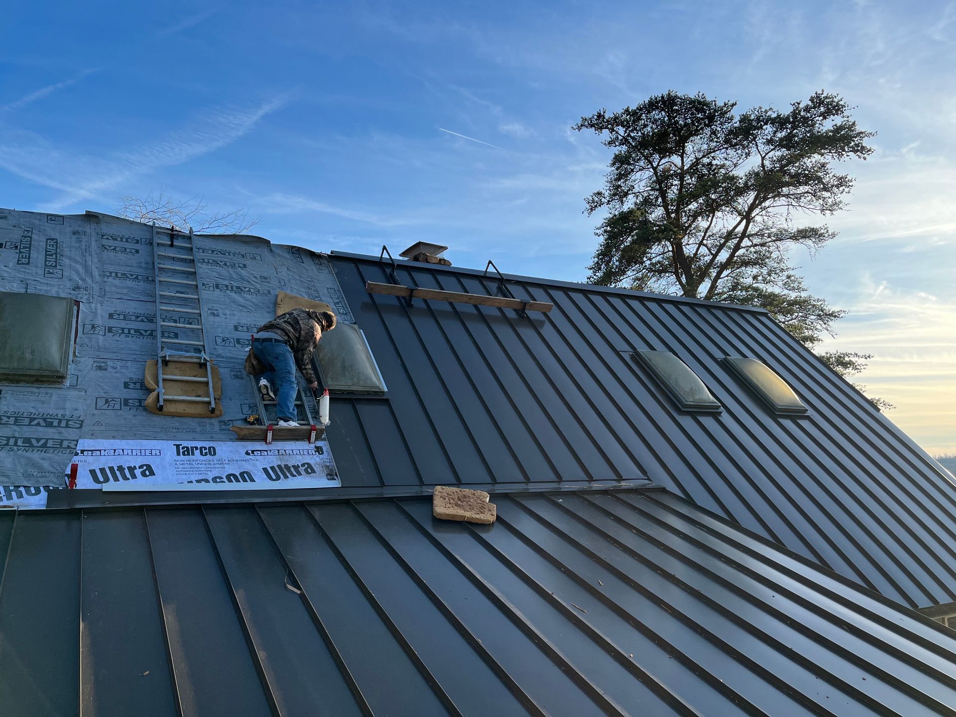 Roofer working on a dark metal roof, installing material, with skylights and ladder visible.