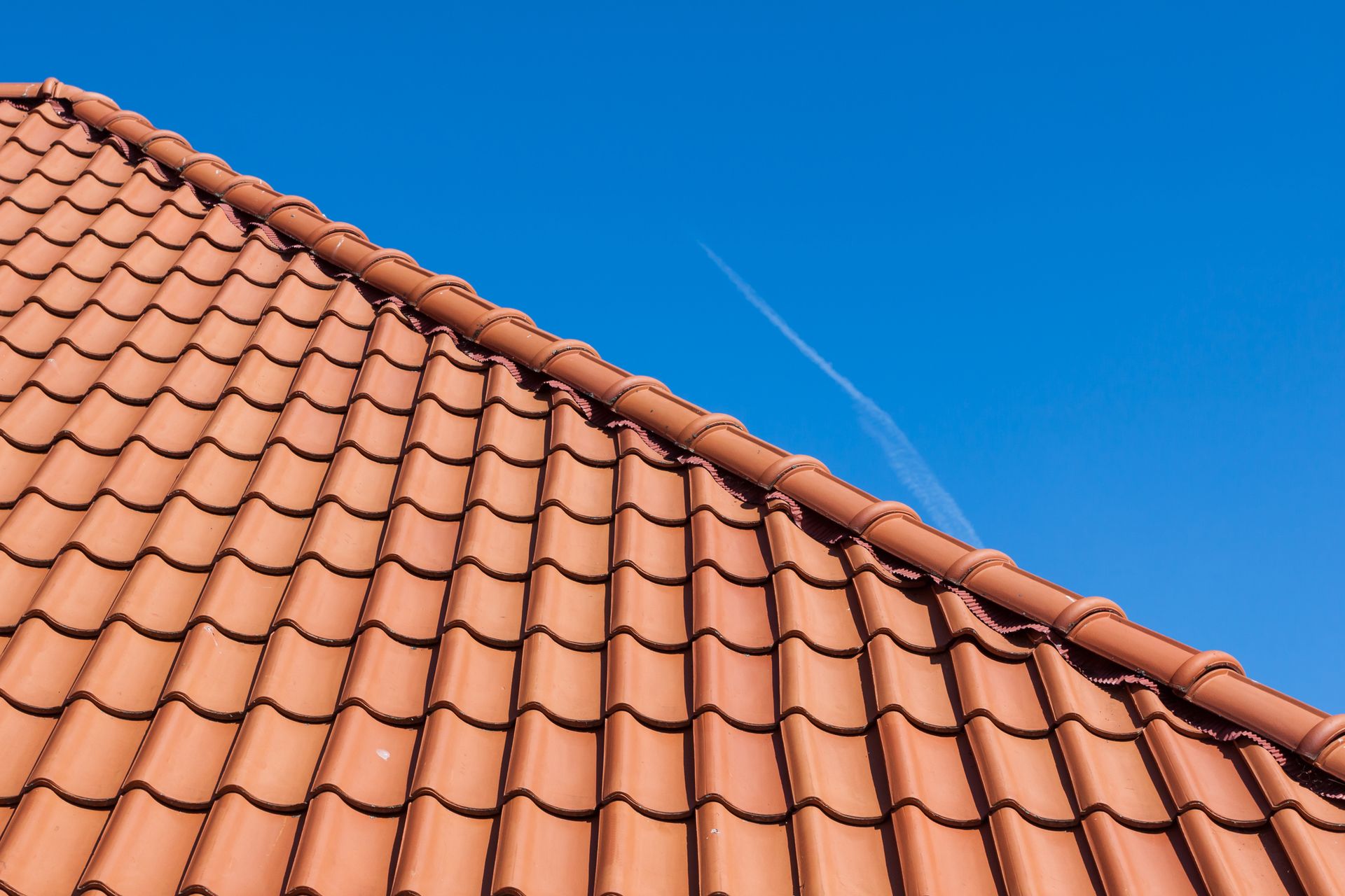 Red clay tile roof against a clear blue sky, with a contrail.