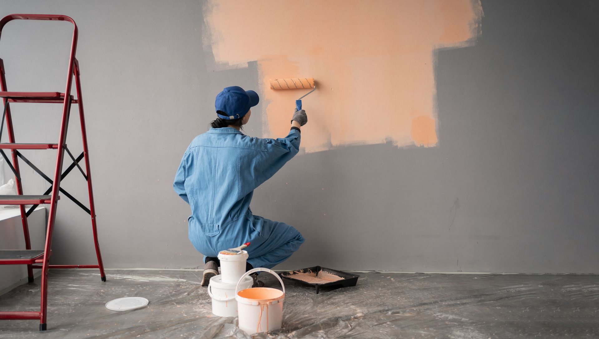 Person in blue jumpsuit painting a peach-colored rectangle on a gray wall, using a roller; stepladder nearby.