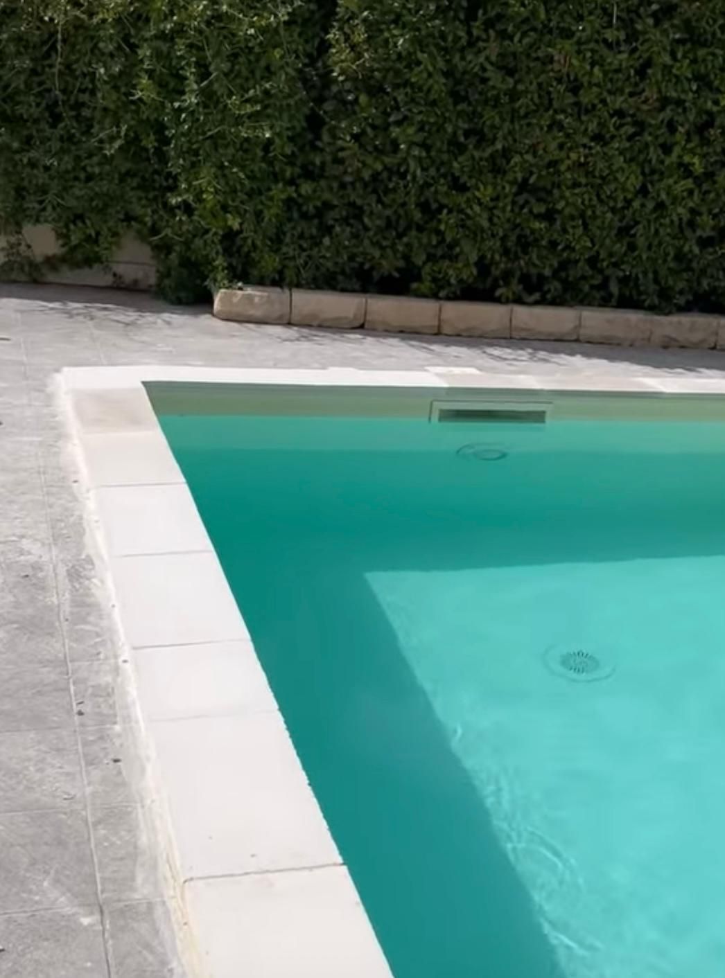 Una piscina con un bordo bianco e una siepe sullo sfondo.