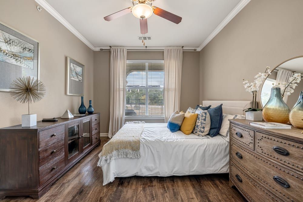 a bedroom with a bed, dresser, mirror, and ceiling fan at Marquis at Crown Ridge in San Antonio, TX.