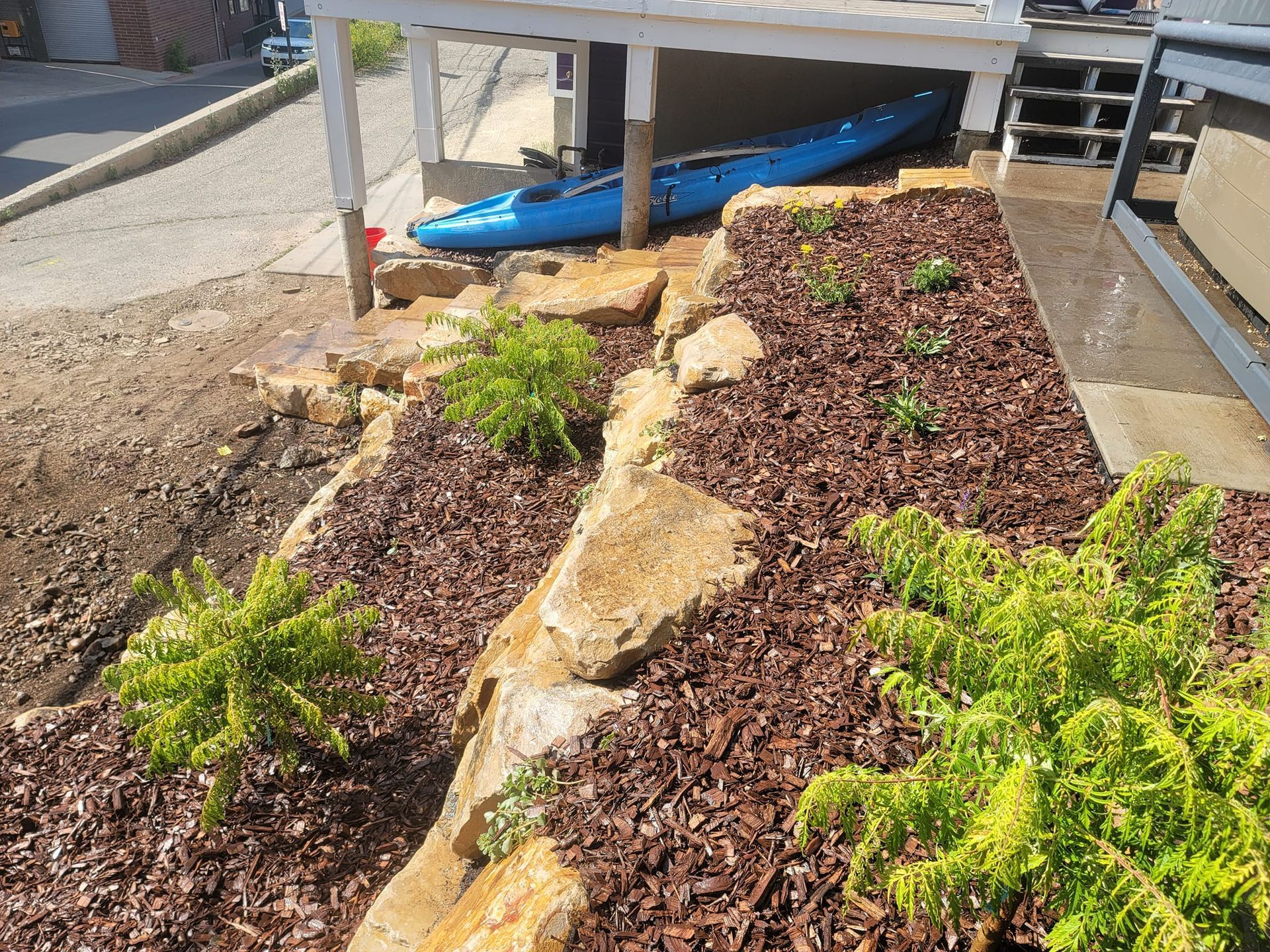 A blue kayak is parked under a deck in a yard.