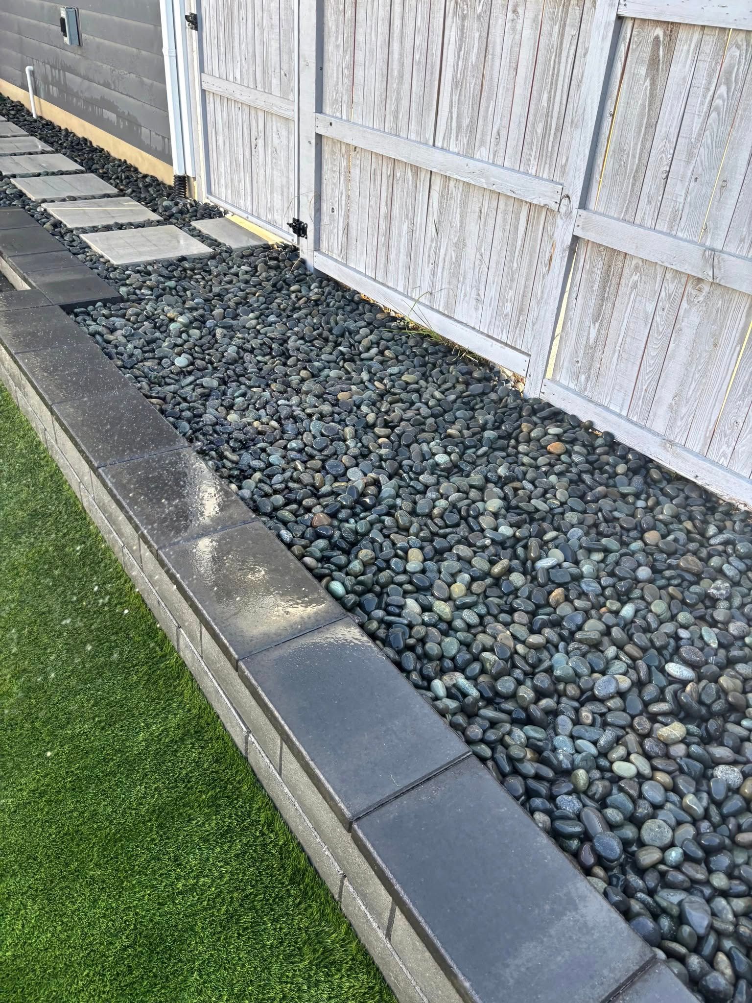 Brick-edged landscaping with green turf, gray stone path, and dark rock bed against a white fence.