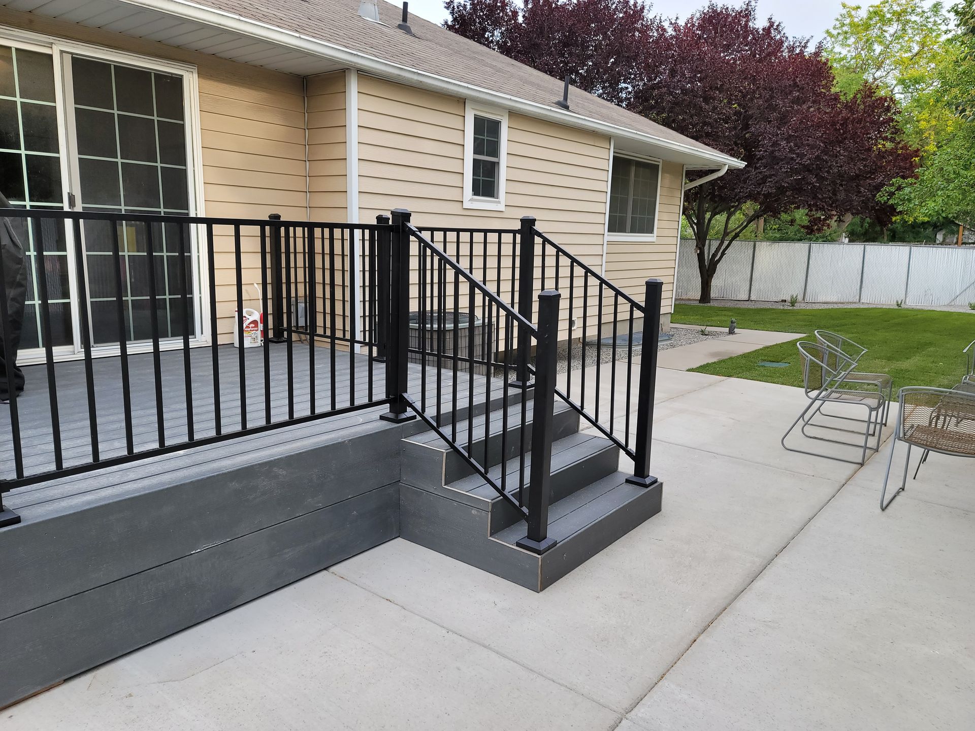 Backyard with deck, stairs, black railings, and concrete patio. House is tan, with green grass and trees.