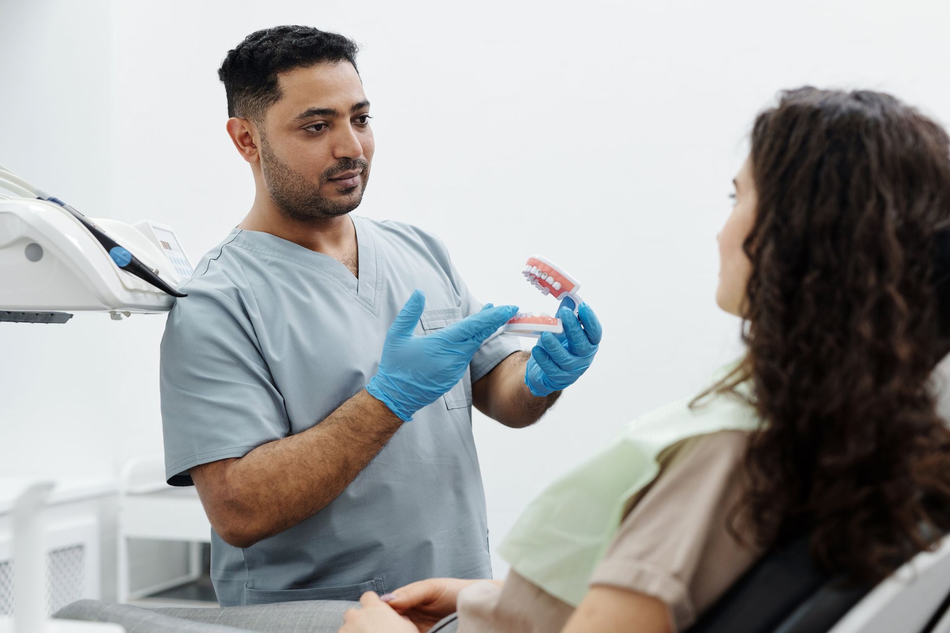 A dentist is talking to a patient in a dental chair while holding a model of teeth.