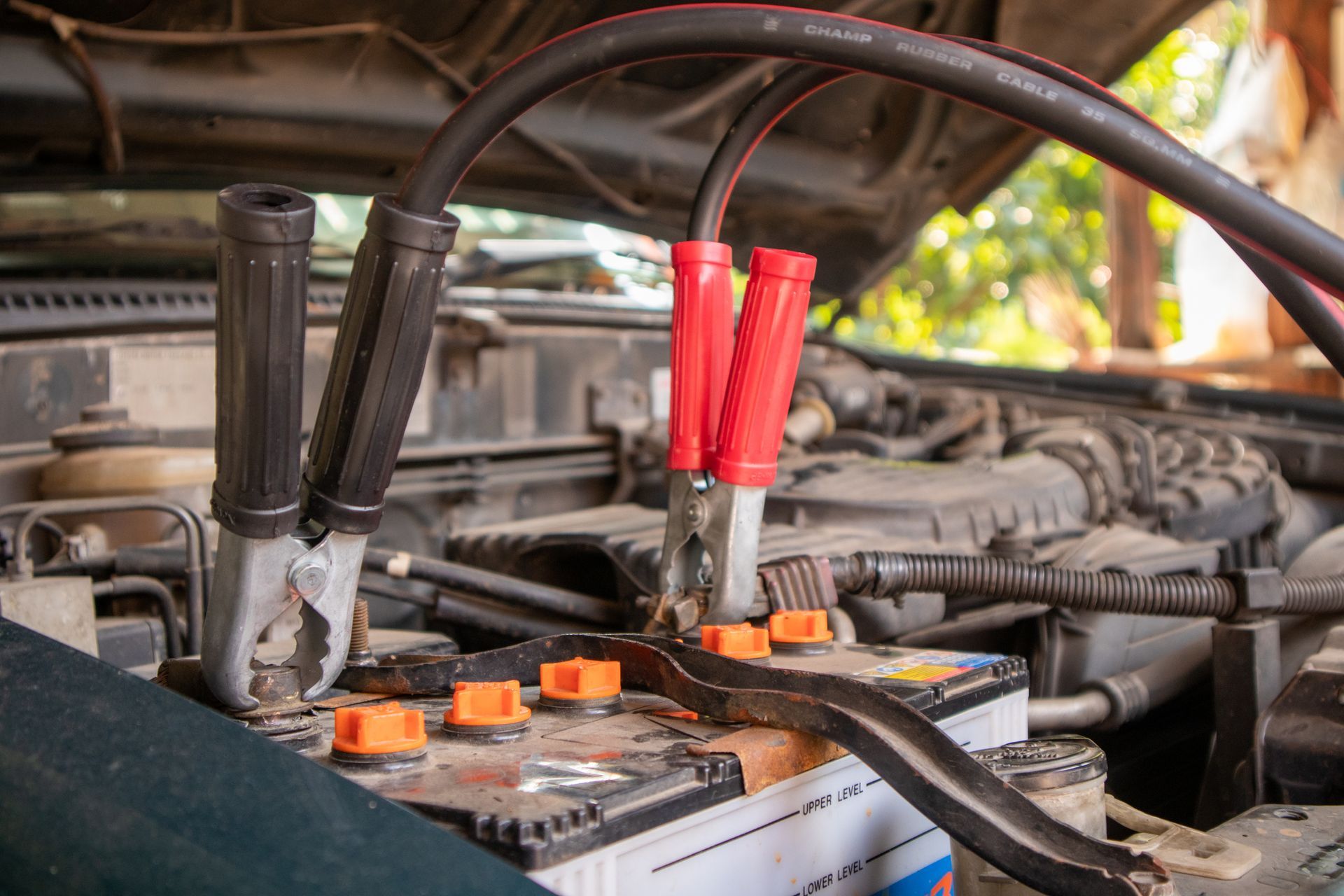 Jumper cables connected to a car battery in an engine bay, the red clamp is attached.