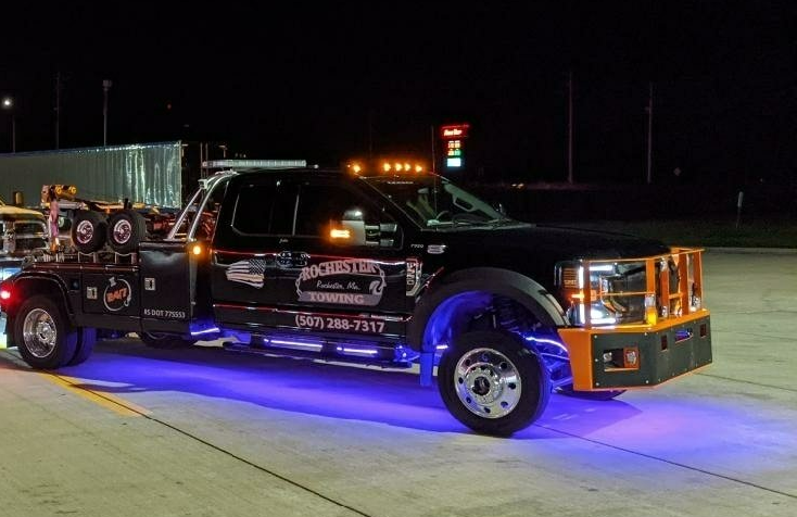 Black tow truck with orange accents and blue underglow lights at night.