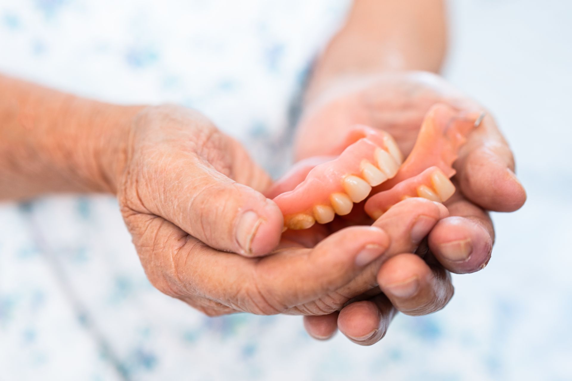 Hands holding a set of upper and lower dentures, pink with white teeth.
