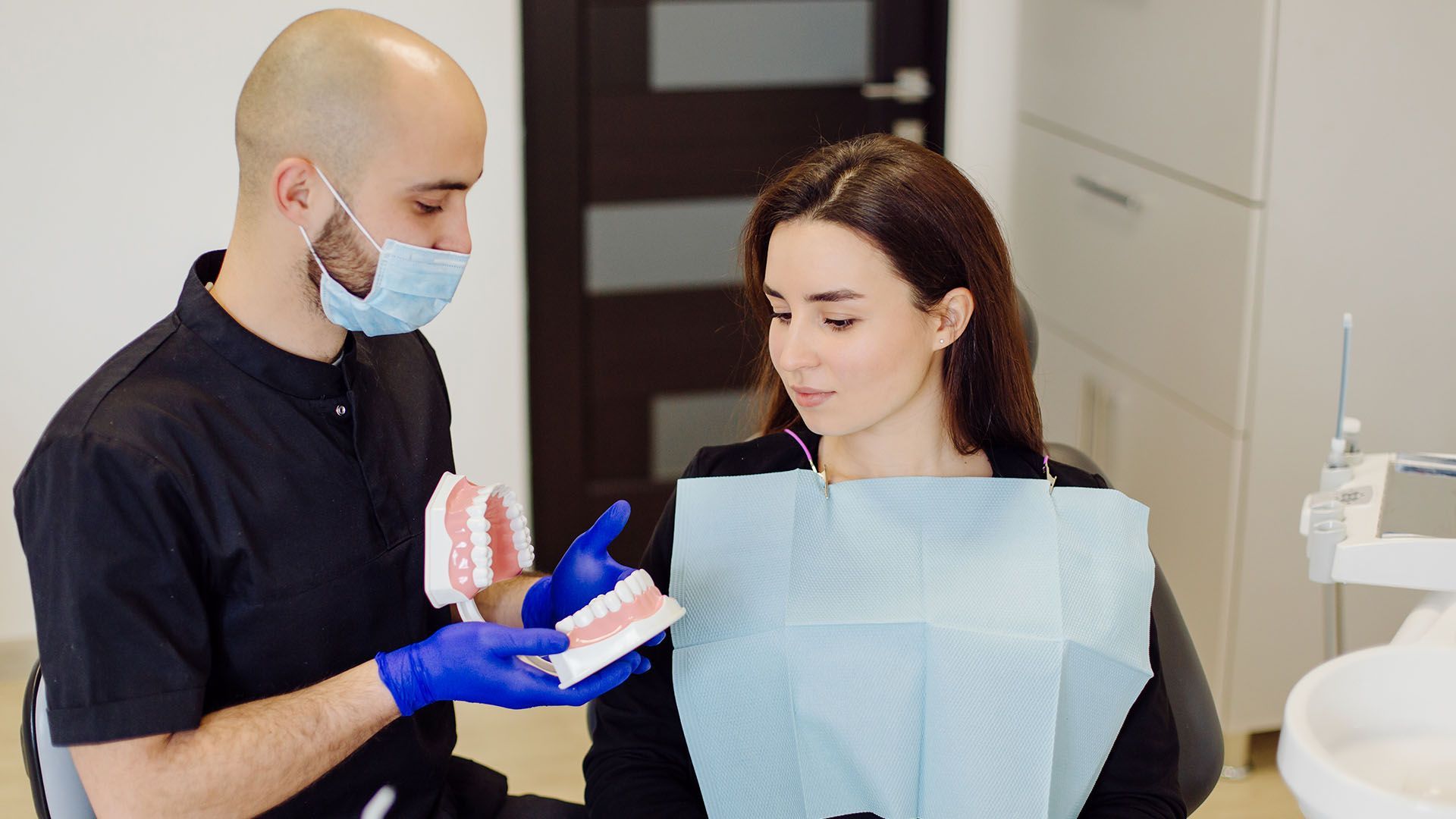 A dentist in a mask and gloves shows a teeth model to a woman in a dentist's chair.