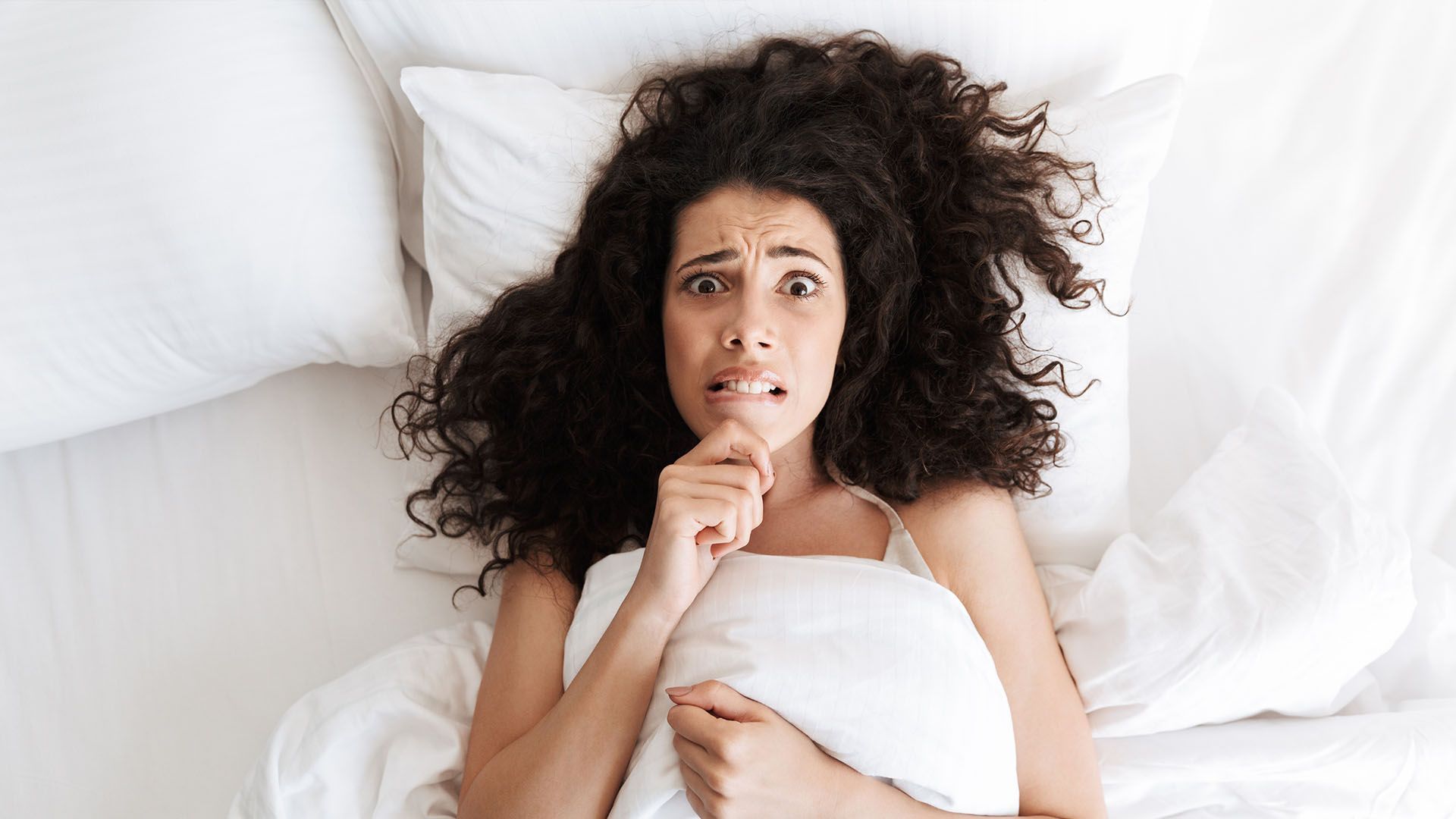 Woman with curly hair in bed, looking frightened and clutching a pillow. White bedding.