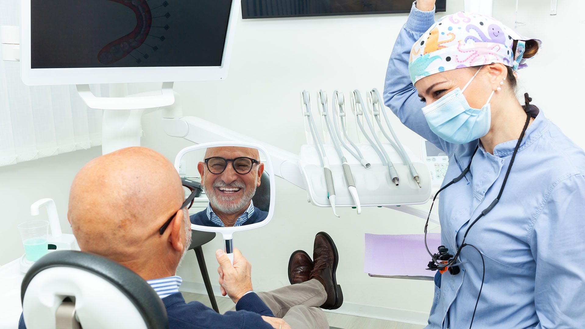 Dentist showing a smiling older man his teeth in a mirror. Both are in a dental office, the dentist is wearing a mask and surgical cap.