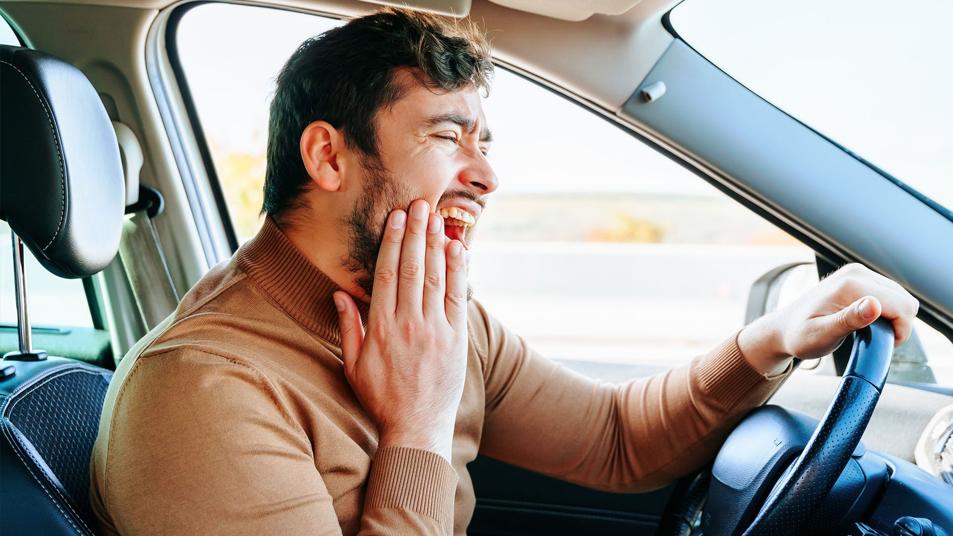 Man driving a car, grimacing and clutching his jaw in pain. Inside car, daytime.