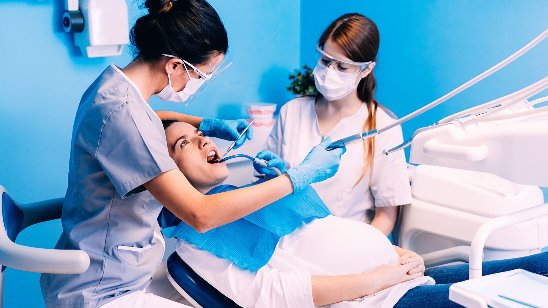 Pregnant woman at the dentist with two dental professionals examining her mouth. Blue-toned room with dental equipment.
