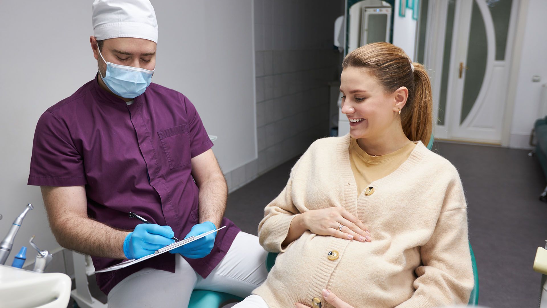 A pregnant woman smiles while sitting in a dental chair, talking to a dentist