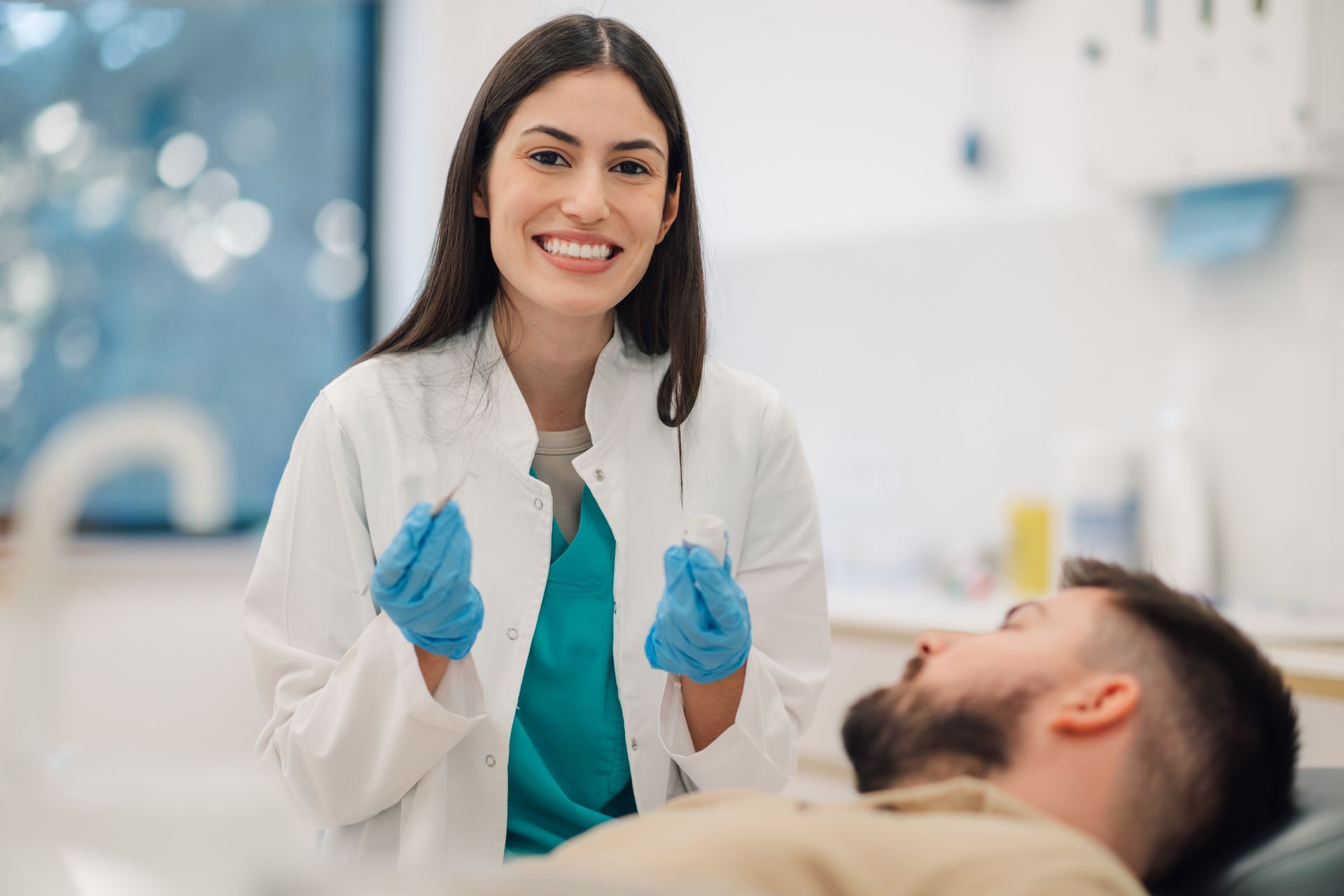 A professional in a white coat and blue gloves smiles while holding dental tools over a patient in a medical setting.