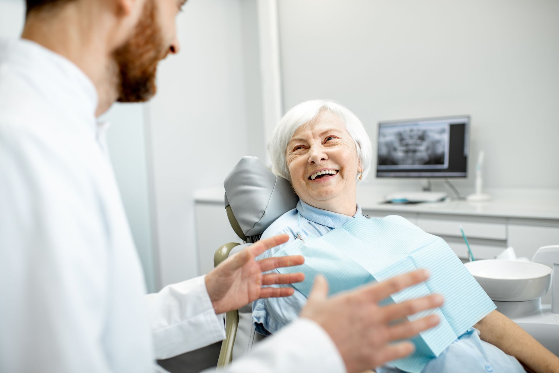 Dentist talking to a smiling elderly woman in a dental chair. X-ray monitor in background.
