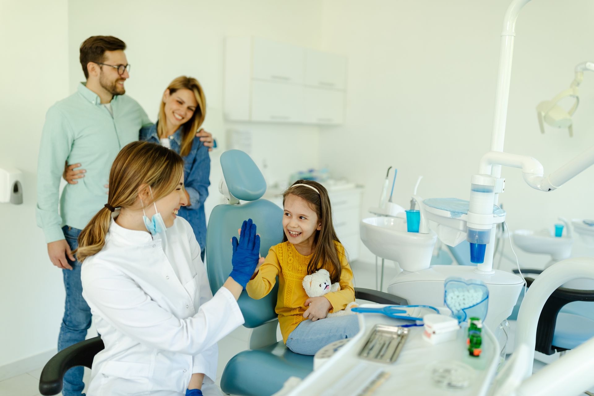 Dentist high-fives a young girl in dental chair; parents watch in bright, modern office.