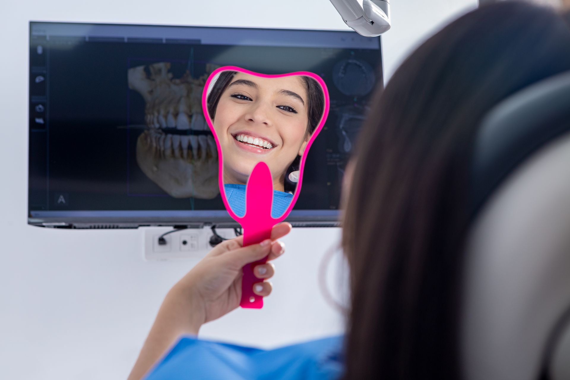 Woman looking at her teeth in a pink mirror at the dentist. A panoramic x-ray image is on the monitor.