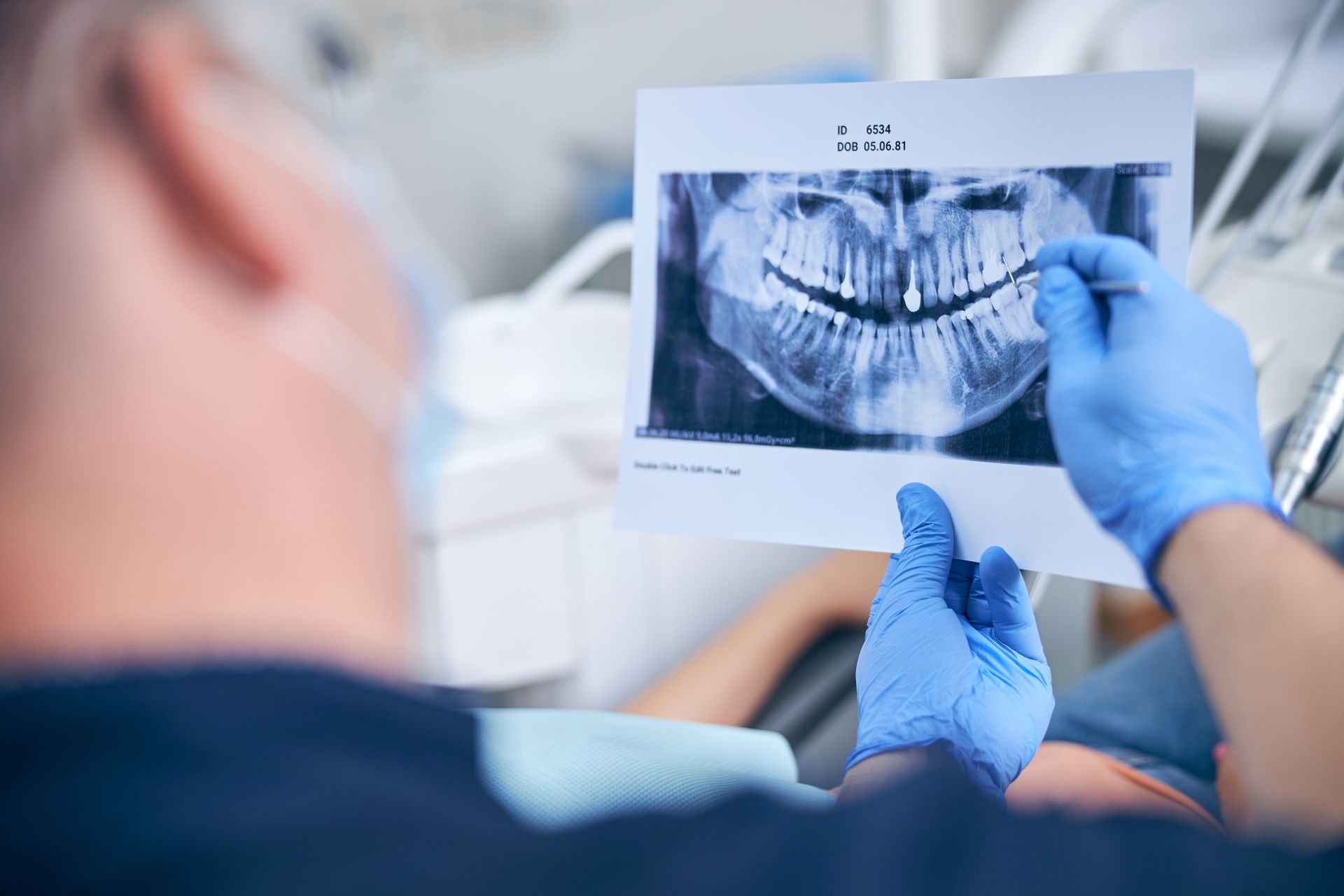 A dentist wearing blue gloves holds and points to a panoramic dental X-ray while consulting with a patient in an office.