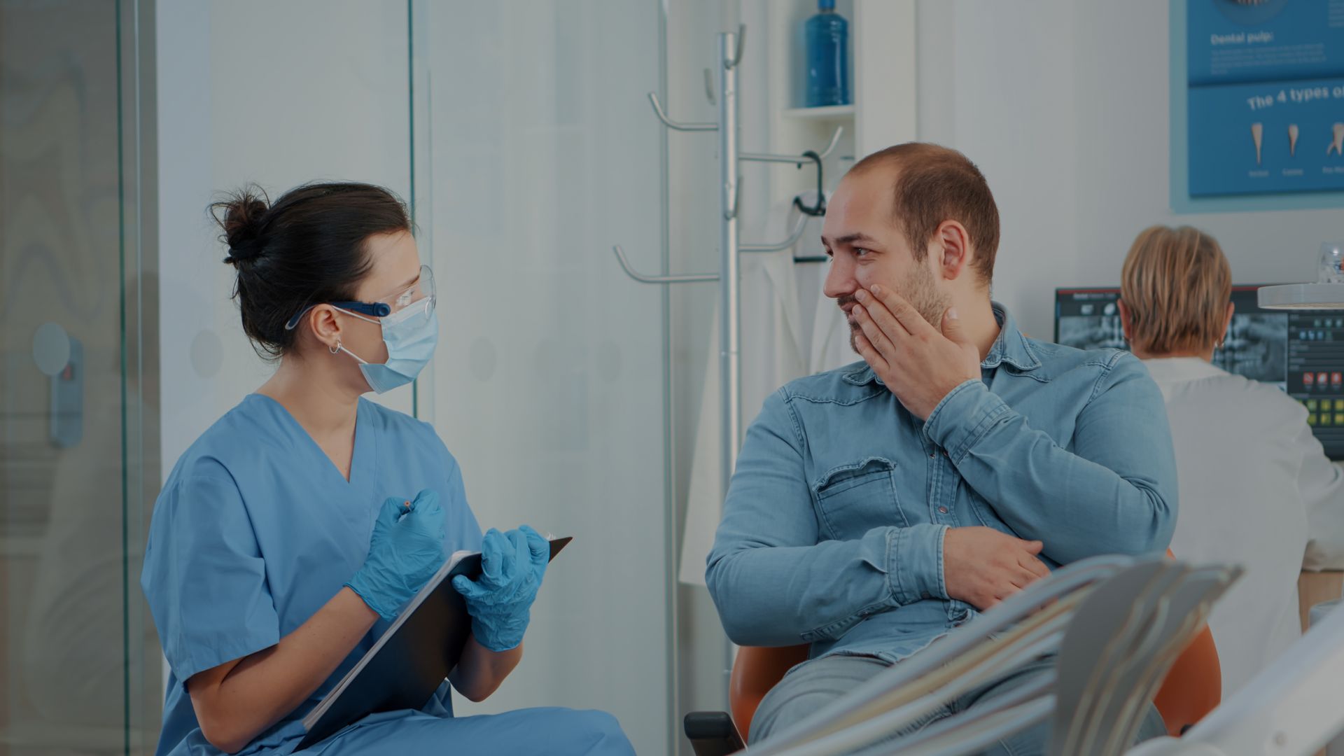Dental assistant in scrubs consults patient with hand on face in dental office.