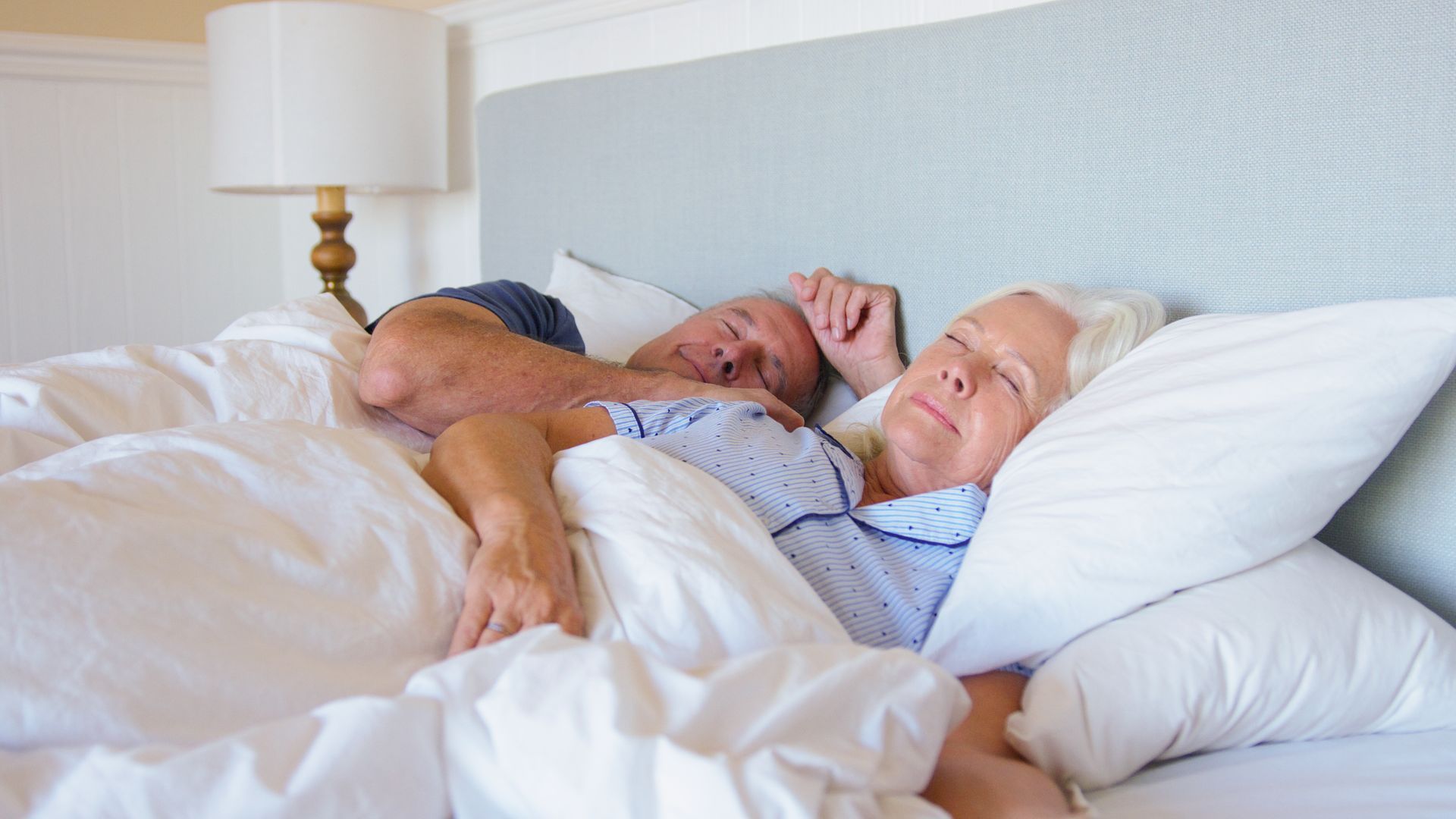Couple sleeping in bed, white bedding, woman smiling, lamp in background.