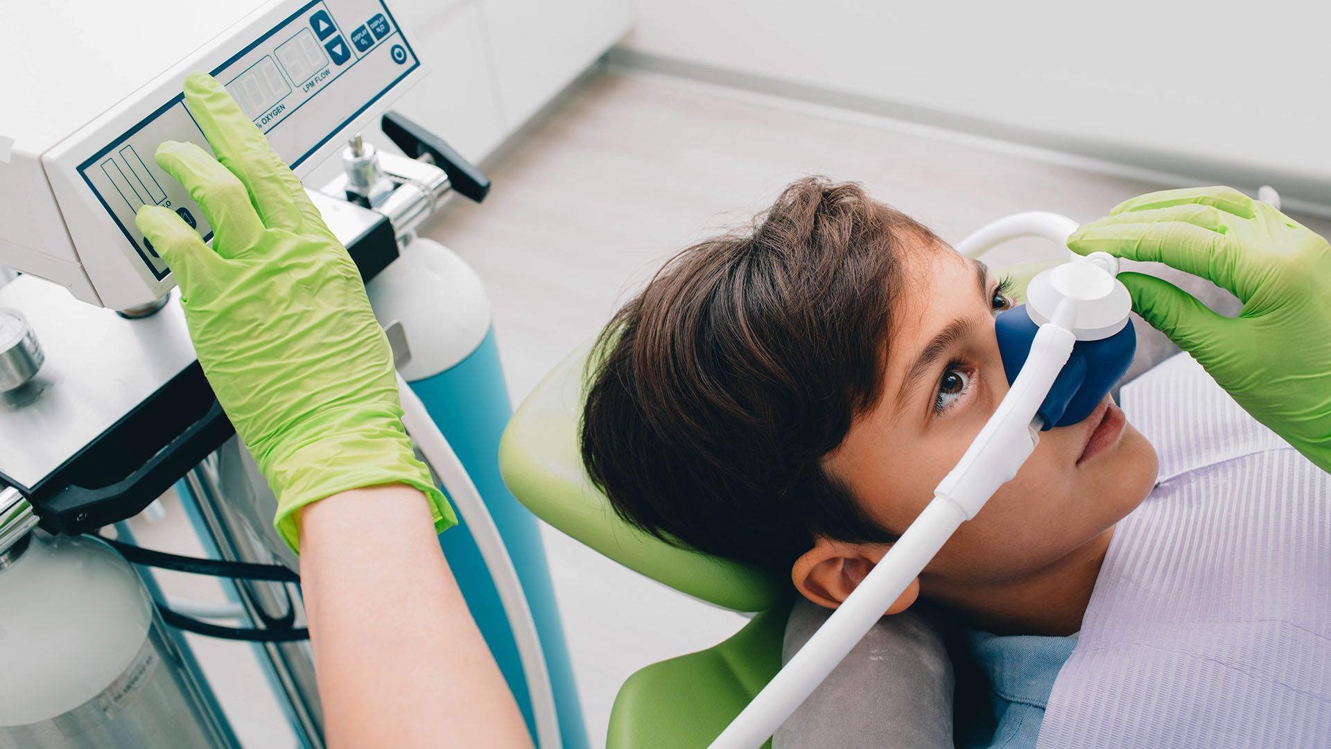 Child receiving dental care with a mask; a dentist's gloved hand adjusts equipment.