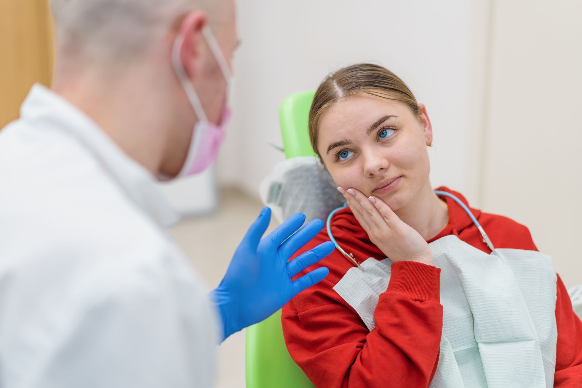 Dentist with patient, patient holding jaw, wearing bib, red sweatshirt. Dentist wears mask, blue gloves.
