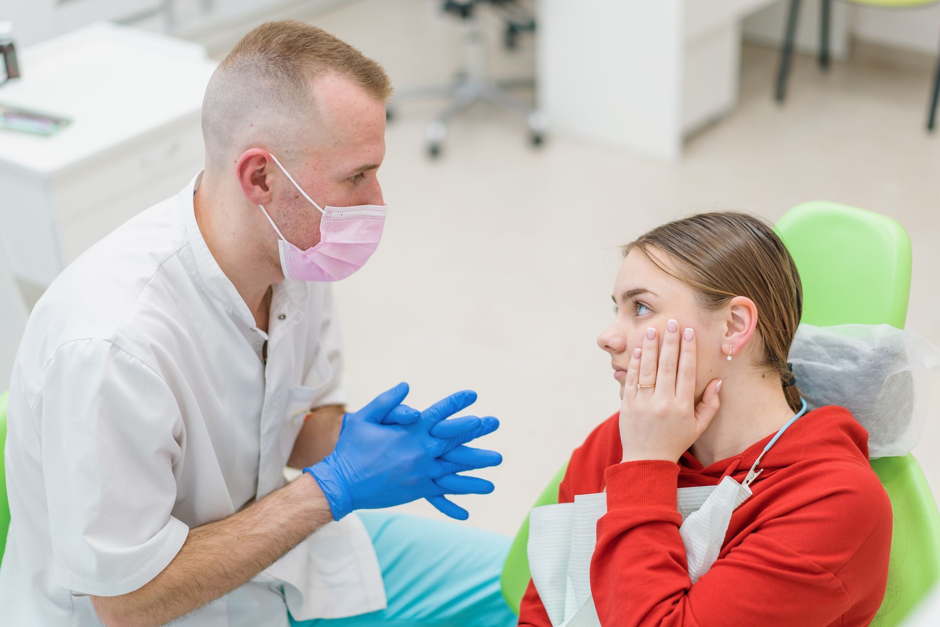 Dentist in mask and gloves talking to a patient holding her cheek, in a dental office.