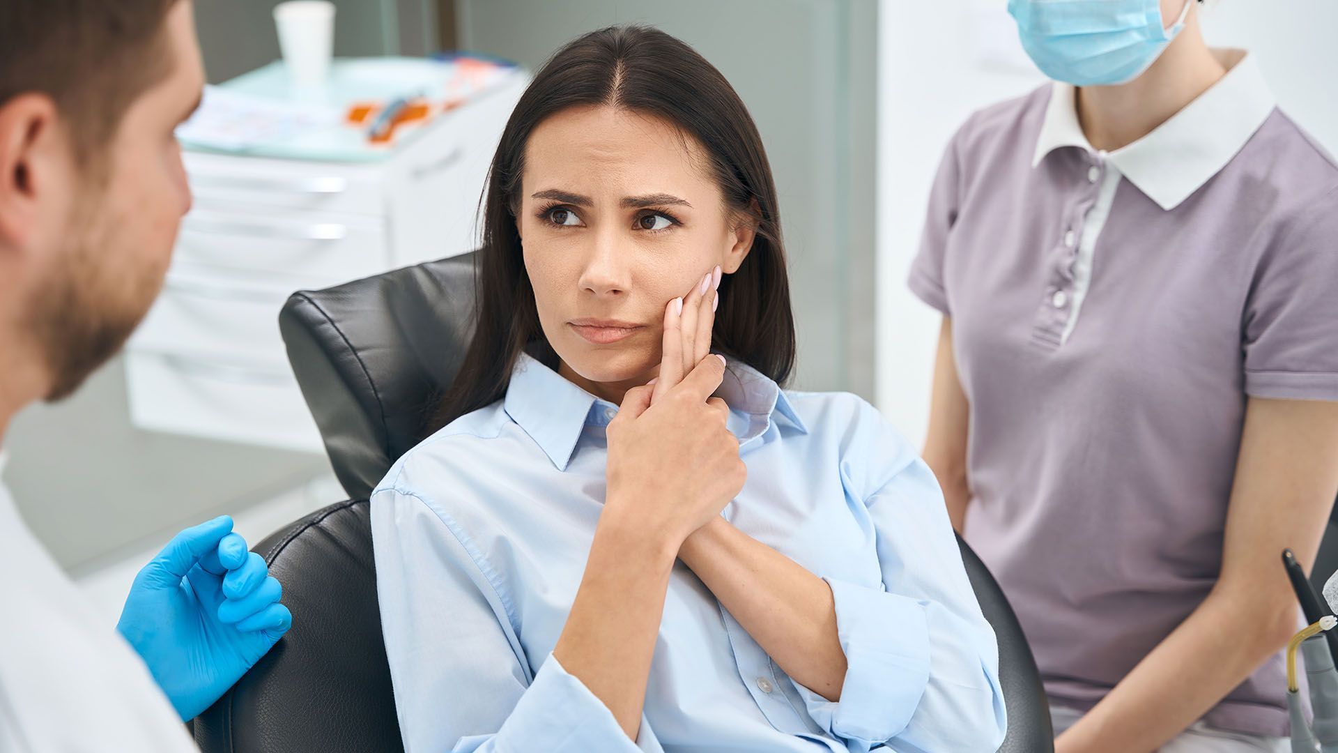 Woman in dental chair, holding jaw, appearing distressed; dentist and assistant in background.